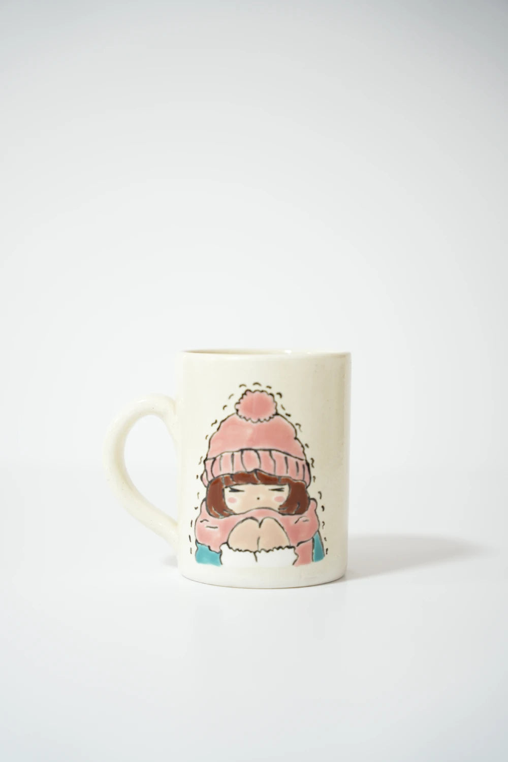 فنجان سيراميك أبيض مع رسمة بنت كرتونية ترتدي قبعة شتوية وردية وجاكيت سميك، جالسة وملتفّة بملابسها وكأنها تشعر بالبرد.