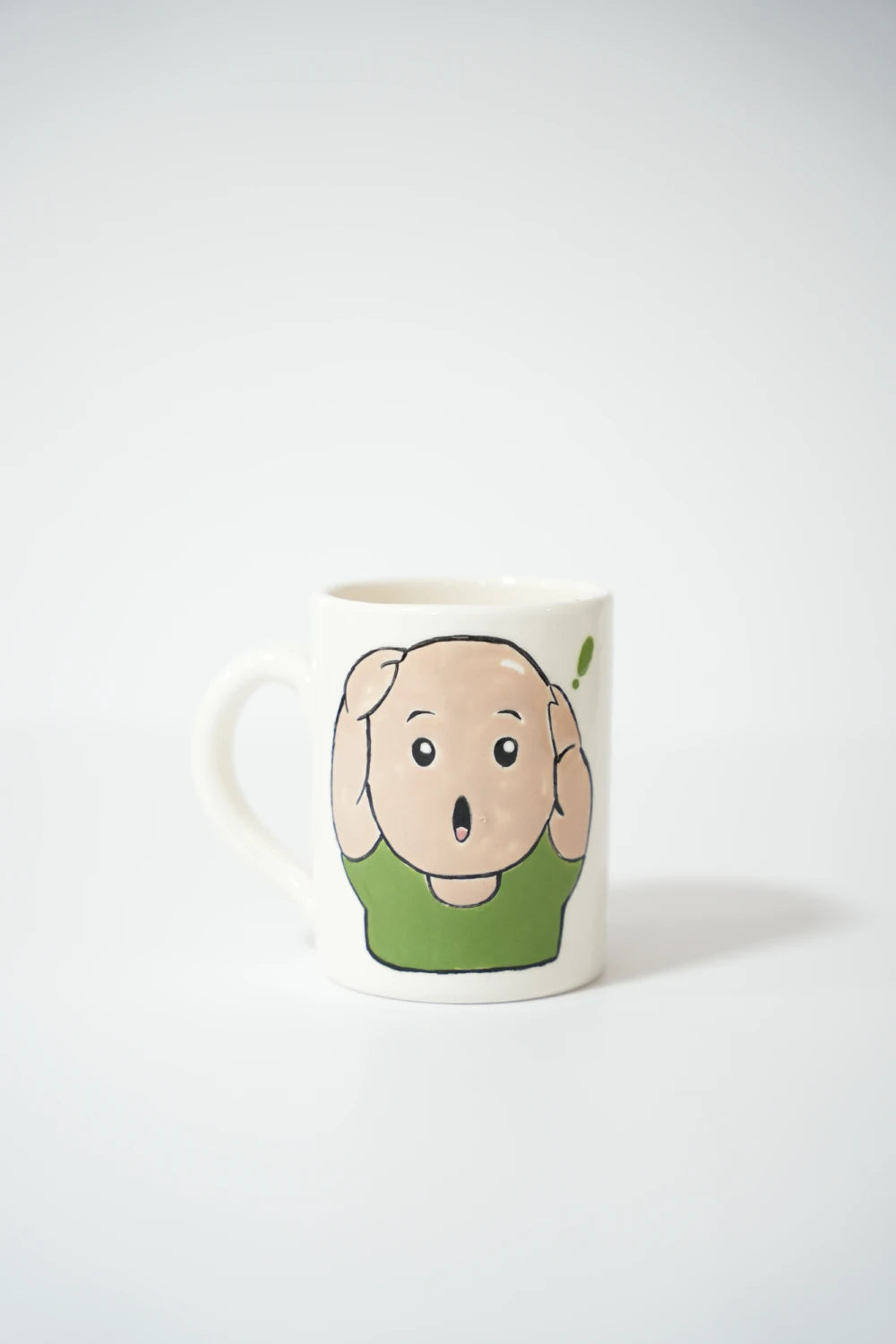 فنجان سيراميك أبيض مع رسمة شب أصلع كرتوني يرتدي بلوزة خضراء، يضع يديه على رأسه بتعبير مصدوم وفم مفتوح.