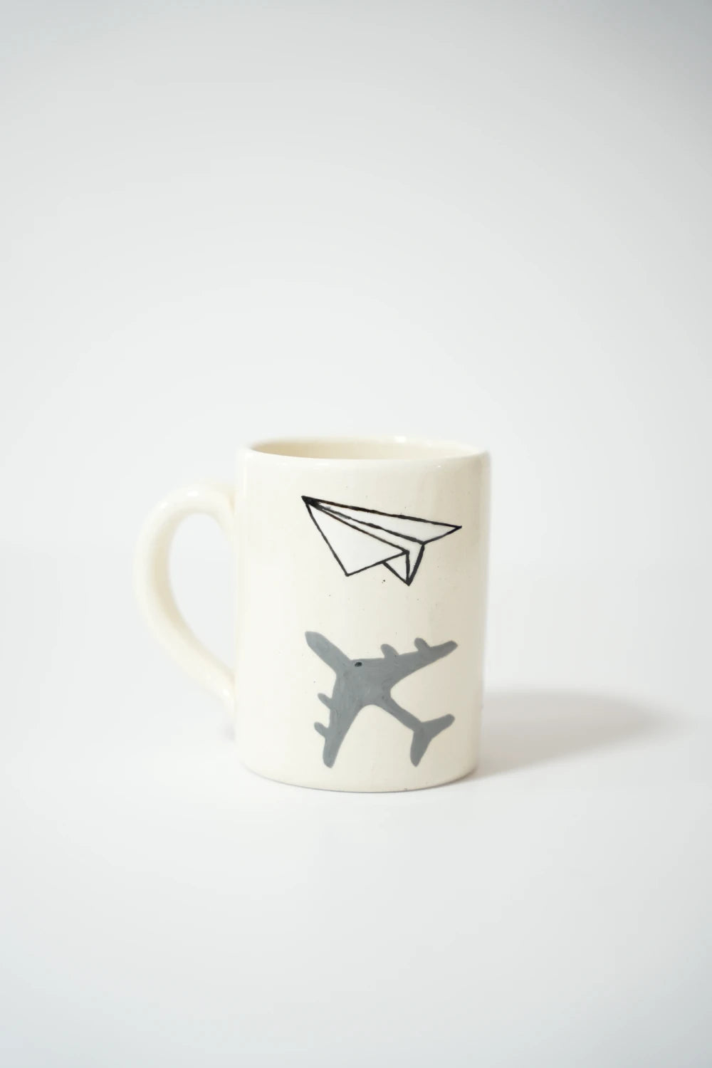 فنجان سيراميك أبيض مرسوم عليه طائرة رمادية كبيرة من الأسفل وطيّارة ورقية مرسومة بخط أسود بسيط في الأعلى، على خلفية نظيفة.