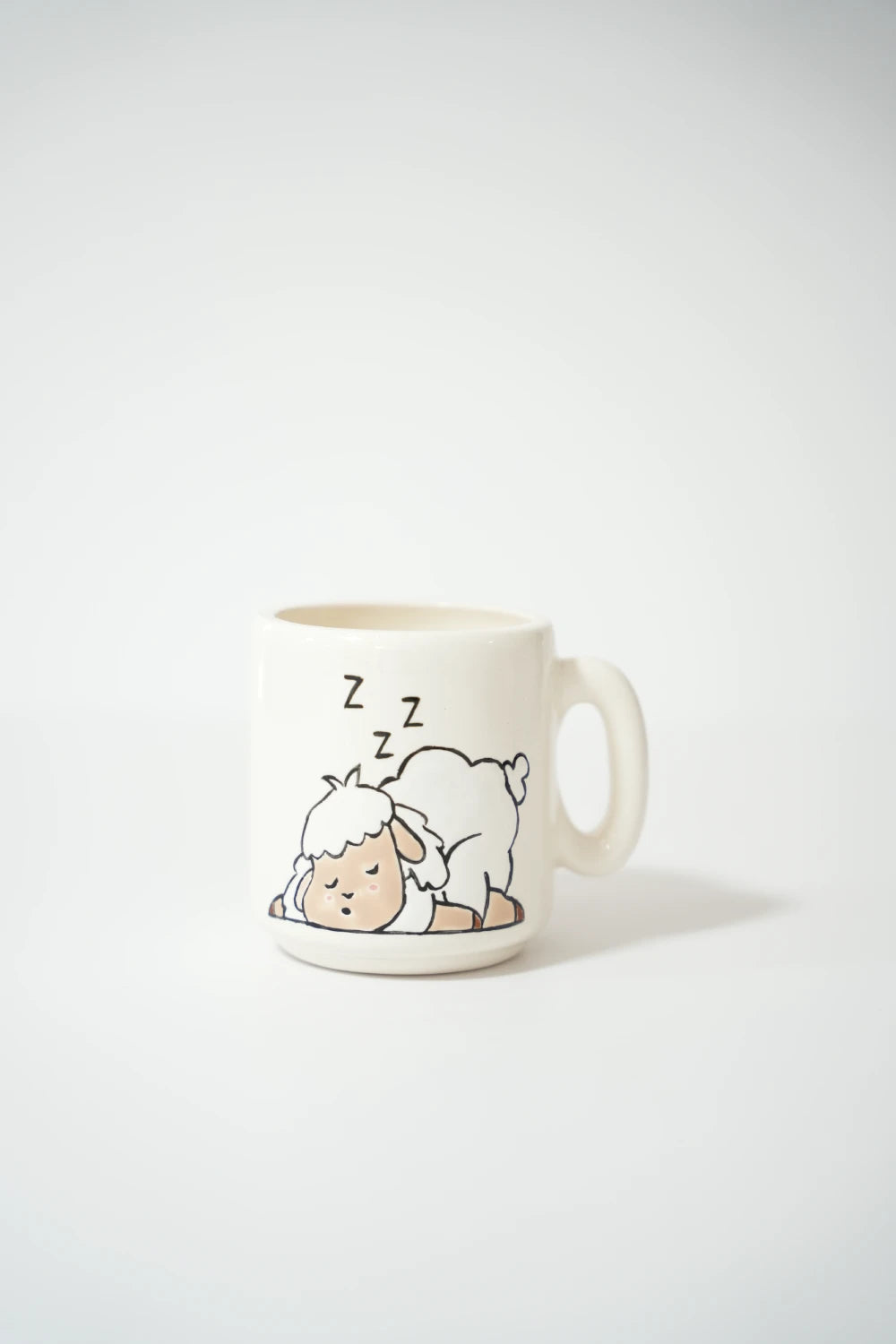 فنجان سيراميك أبيض مرسوم عليه خروف كرتوني صغير نايم على الأرض، مغمض عينيه مع رموز Zzz فوق رأسه في وضعية نوم لطيفة.