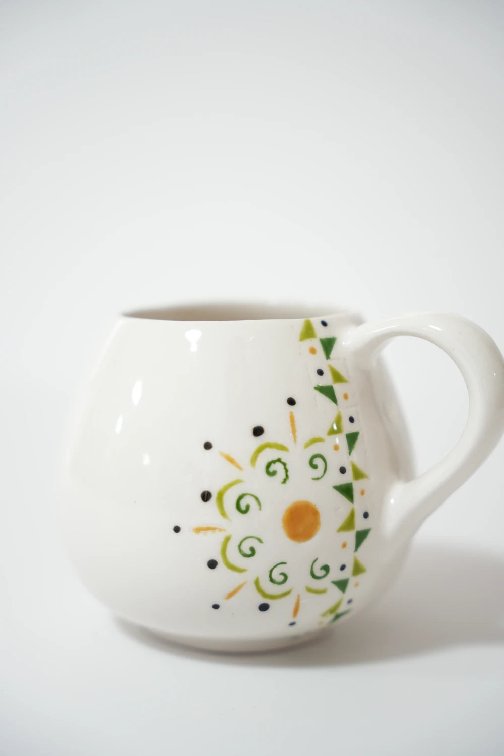 فنجان سيراميك أبيض دائري الشكل بنقوش يدوية على الجهة الجانبية، عبارة عن دائرة زخرفية بلون برتقالي في المنتصف مع لفّات خضراء ونقاط سوداء، وبجانبها صف مثلثات خضراء صغيرة بشكل زخرفة شرقية.