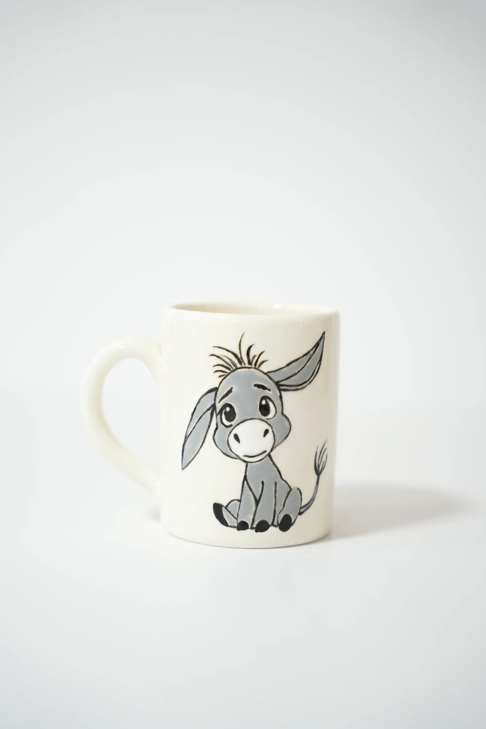 فنجان سيراميك أبيض مرسوم عليه حمار كرتوني رمادي يجلس على الأرض، بأذنين طويلتين وعيون كبيرة لطيفة وتعبير بسيط يميل للتعب والبراءة.