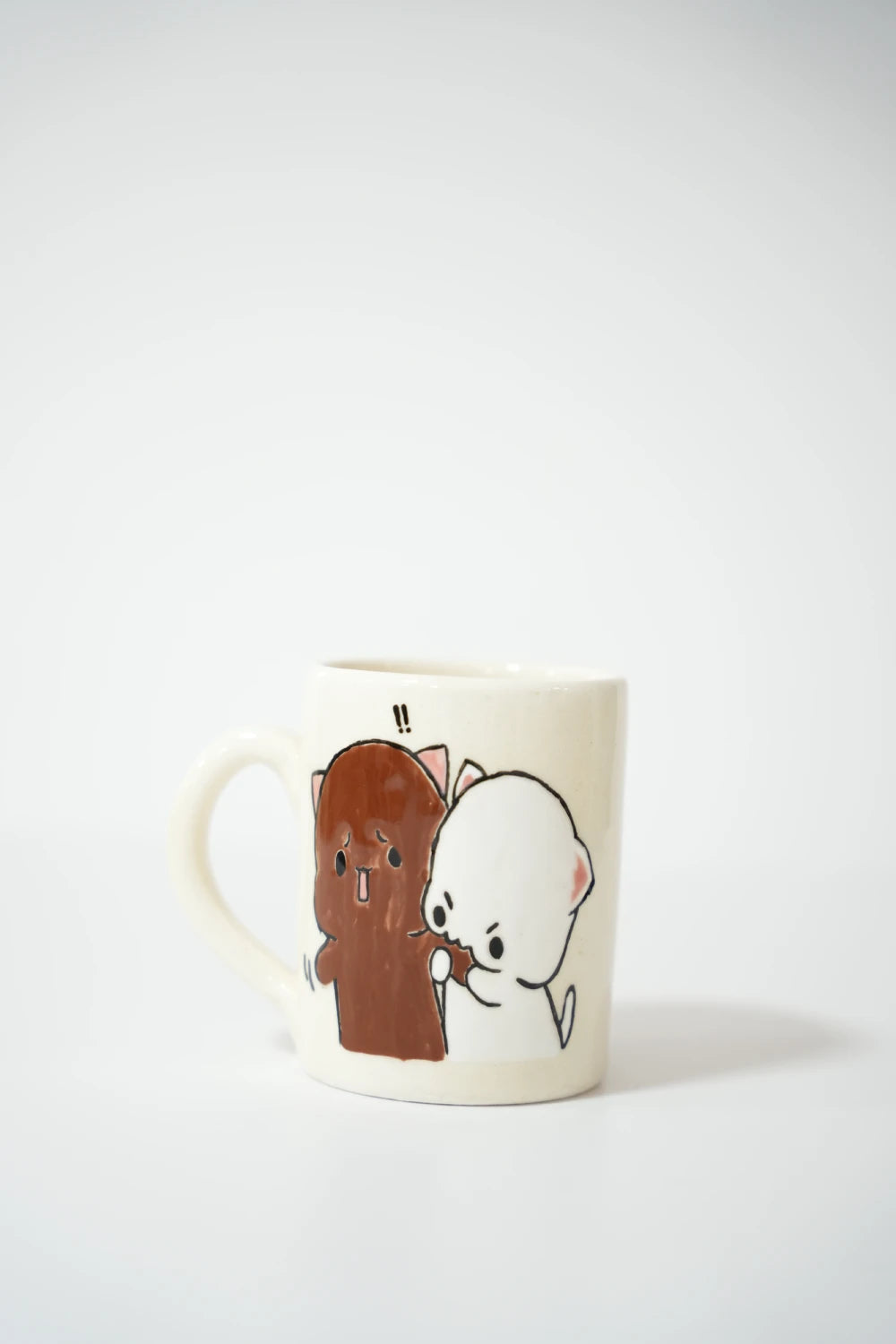 فنجان سيراميك أبيض مرسوم عليه شخصيتان كرتونيتان لطيفتان تشبهان القطط أو الدببة الصغيرة، إحداهما بنية اللون بملامح مندهشة، والأخرى بيضاء تقف خلفها وتمسك بها مع علامتي تعجب فوقهما.