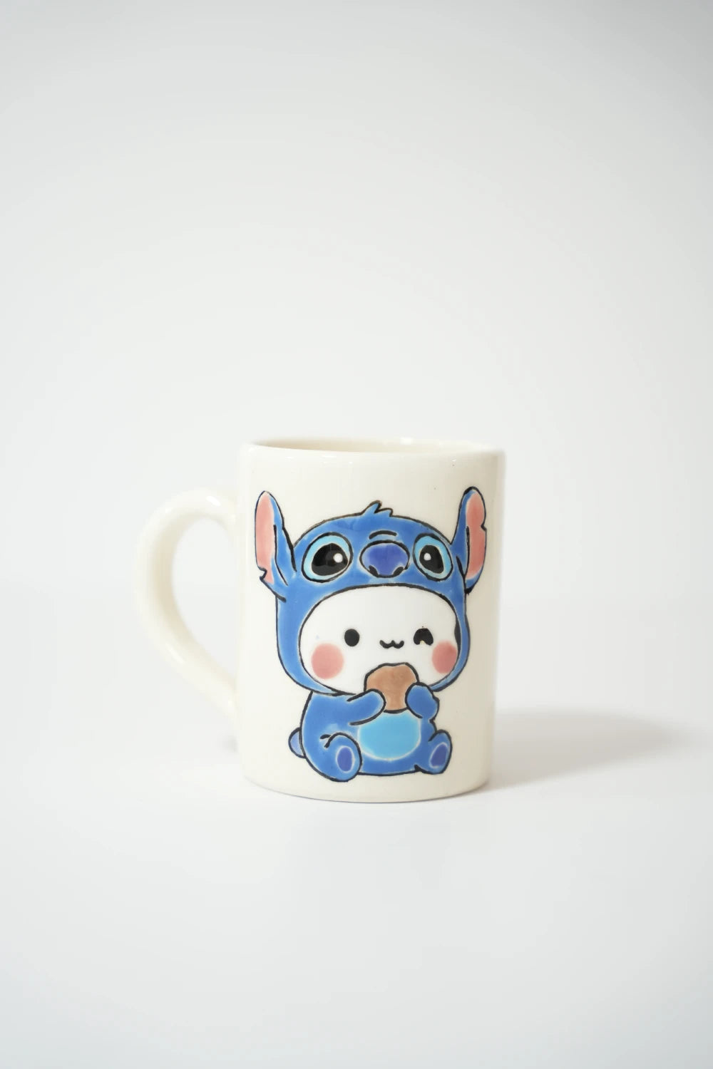 فنجان سيراميك أبيض مرسوم عليه شخصية كرتونية صغيرة ترتدي بدلة زرقاء كاملة مع أذنين كبيرتين، تجلس وتمسك قطعة بسكويت بيديها ووجنتيها ورديتان على خلفية بيضاء بسيطة.