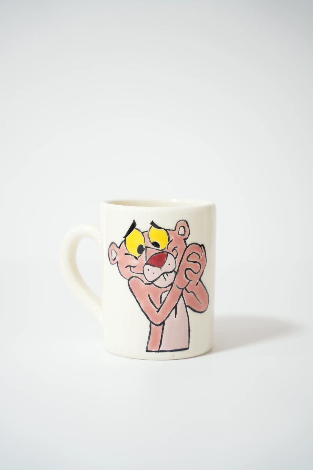 فنجان سيراميك أبيض مرسوم عليه النمر الوردي الكرتوني بتعبير متمنّي ويديه مضمومتين.