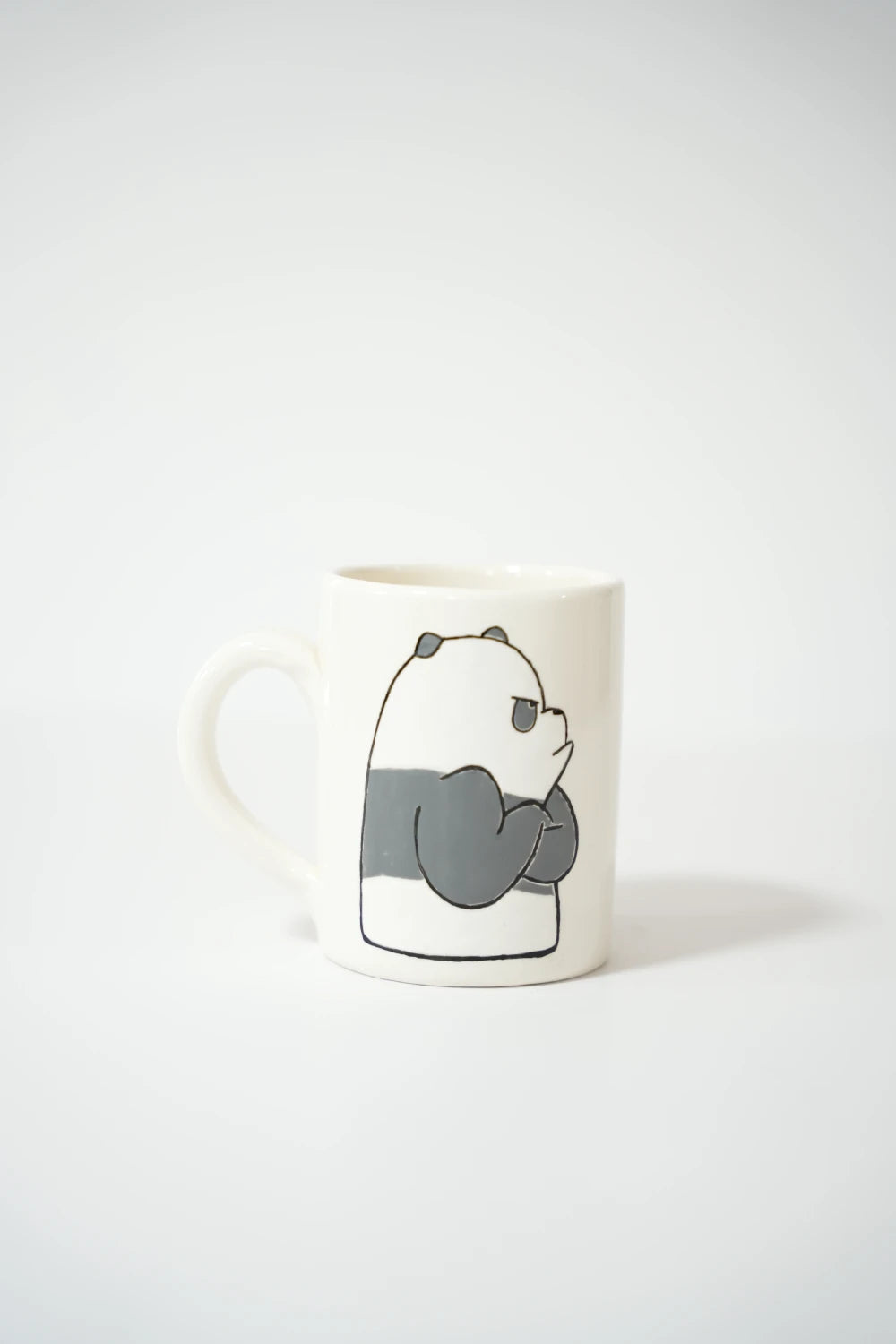 فنجان سيراميك أبيض عليه رسمة باندا كرتونية باللونين الرمادي والأبيض، حاطّة إيديها ومبيّنة تعبير زعل أو انزعاج.